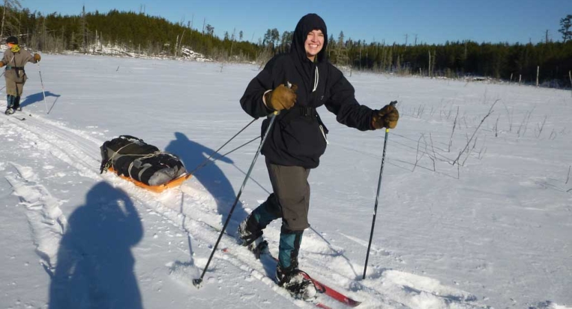 A person smiles as they cross country ski across a vast snowy landscape, pulling a small sled behind them. 